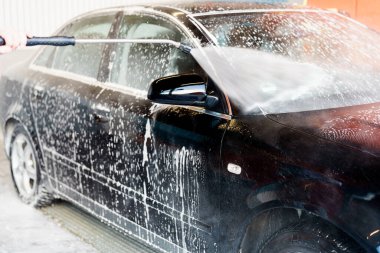 Birisi araba yıkamada köpükle kaplı siyah bir arabayı yıkamak için yüksek basınçlı sprey kullanıyor. Tesis aydınlık ve hareketlilikle dolup taşıyor. Güneşli bir öğleden sonra..