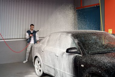 Bir adam, kapalı araba yıkama yerindeki bir arabaya temizlik solüsyonu uygulamak için köpük topu kullanıyor. Köpük aracı kaplıyor, temizlik yaparken parlak bir görünüm yaratıyor..