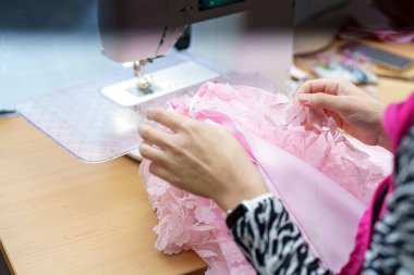 Bir insan, dikiş makinesinde pembe dantel kumaşı dikkatlice diker, karmaşık tasarım becerileri sergiler ve el işi aletleriyle dolu parlak bir çalışma alanında işçiliğe odaklanır..