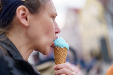Neşeli bir kadın, güzel ve hareketli bir atmosferle çevriliyken mavi bir dondurma külahının tadını çıkarıyor. Sahne, güneşli bir günün neşesini ve sıcaklığını yakalar..