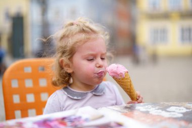 Küçük bir kız masada oturmuş pembe bir dondurma külahının tadını çıkarıyor. Yüzündeki neşeli ifade, bir ısırık aldığında verdiği hazzı gösteriyor. Mekan canlı bir meydan..