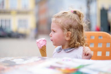 Kıvırcık saçlı genç bir kız parlak turuncu bir masada pembe dondurma külahının tadını çıkarıyor. Arka plan bulanık binaları içeriyor, canlı bir açık hava ortamını gösteriyor.