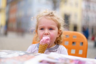 Kıvırcık saçlı genç bir kız renkli bir kafe masasında oturmuş pembe dondurma külahının tadını çıkarıyor. Çevre şehir meydanı hareketli, günün tadını çıkaran insanlarla dolu..
