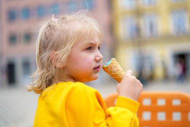 Açık renk saçlı bir çocuk açık havada otururken dondurma külahını ısırıyor. Çevredeki renkli binalar neşeli bir atmosfer yaratıyor, sıcak bir gün için mükemmel..