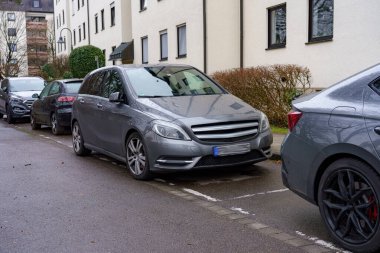 Gri bir araç başka bir arabanın yanına park edilmiş dar bir caddede konut binalarıyla dolu. Hava bulutlu görünüyor, serin bir atmosfer olduğunu gösteriyor..