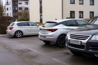 Modern binalarla çevrili bir konutun otoparkına birkaç araba park edilmiş. Hava bulutlu, bölgede bastırılmış bir atmosfer yaratıyor..