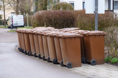Çok sayıda kahverengi çöp tenekesi sakin bir banliyö bölgesinde kaldırım kenarı boyunca düzgünce düzenlenir ve gündüzleri yerleşim yerlerinde atık yönetimi uygulamaları sergilenir..