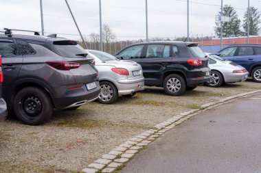 Bir spor tesisine bitişik çakıllı bir otoparkta çeşitli araçlar var. Sahnede bulutlu gökyüzü altında çeşitli araba modelleri ve renkler yer alıyor..