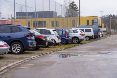 Çeşitli araçlar bir spor tesisinin yanındaki çakıllı yolda düzgün bir şekilde park edilmiş. Gökyüzü bulutlu ve alan çimen ve çitle çevrili görünüyor..