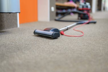 Elektrikli süpürge halı kaplı bir zeminde oturuyor, ofis ortamında kullanılmaya hazır. Çalışma alanı düzenli görünüyor, verimlilik için temizliğin önemini vurguluyor.