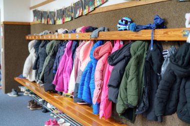 Bir okul koridorundaki ahşap raflara parlak renklerde çeşitli kışlık paltolar asılıdır. Ayakkabılar aşağıda dizilmiş, canlı bir sabah atmosferi var..