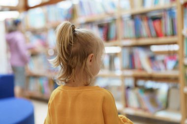Sarı saçlı küçük bir çocuk kütüphanede çeşitli kitaplarla dolu bir kitaplığa bakar. Başka bir kişi daha yakınlarda gezinerek okuma ve keşfetme için huzurlu bir ortam yaratıyor..
