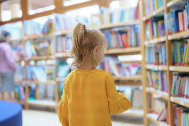 Canlı bir kütüphanede, sarı kazaklı genç bir kız renkli başlıklarla dolu raflardaki kitaplara bakıyor. Başka bir müşteri arka planda geziniyor, barışçıl atmosferin tadını çıkarıyor..