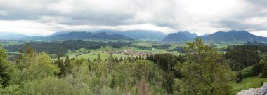 Panoramik bir manzara bulutlu bir gökyüzünün altında canlı yeşil tepeler ve dağlar arasında yuva yapmış sakin bir köy yakalar. Sakin manzara doğanın güzelliğini gözler önüne seriyor..