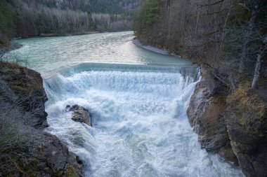 Güçlü bir şelale, yumuşak sabah ışığı altında yemyeşil ağaçlarla çevrili sakin bir nehre dökülür. Huzurlu sahne doğanın güzelliğini yakalar..
