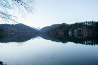 Sakin sular, alacakaranlıkta yakınlardaki dağları ve ağaçları yansıtarak huzurlu ve manzaralı bir atmosfer yaratır. Gökyüzü açık, doğanın güzelliğini arttırıyor..