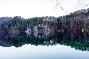 Sabahın erken saatlerinde sakin bir göl üzerinde dingin bir parıltı sarıyor. Yoğun ormanlar ve engebeli uçurumlar ortaya çıkıyor. Durgun su manzarayı çok güzel yansıtıyor. Huzurlu bir atmosfer yaratıyor..