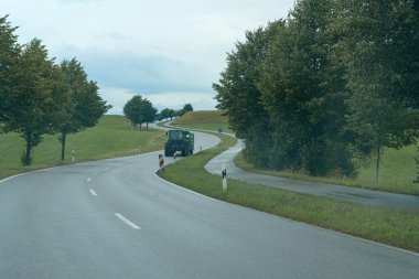Bir traktör, yemyeşil tepeler ve bulutlu bir gökyüzünün altındaki ağaçlarla çevrili dolambaçlı bir yolu takip eder. Sahne, kırsal yaşamda bir huzur hissi yakalıyor..