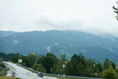 Bir araç virajlı bir yolda ilerlerken sis tepeleri kaplıyor. Yol kenarındaki yemyeşil ağaçlar, dağlardaki bu bulutlu günde sakin bir atmosfer yaratıyor..