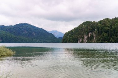 Yeşil tepeler ve gri gökyüzünün altındaki yüksek dağlar arasında sakin bir göl yuva yapar. Yumuşak dalgalar su yüzeyinde dalgalanmalar yaratır ve çevresindeki manzarayı yansıtır..