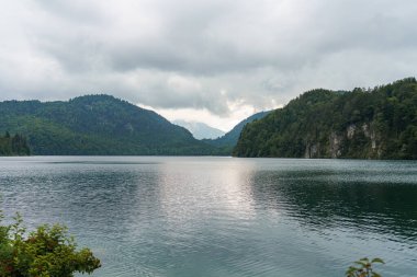 Bulutlar, etrafındaki dağların zengin yeşilliklerini yansıtan sakin bir göl üzerinde duruyor. Durgun su, sabah ışığında doğanın güzelliğini gösteren huzurlu bir kaçış sunar..