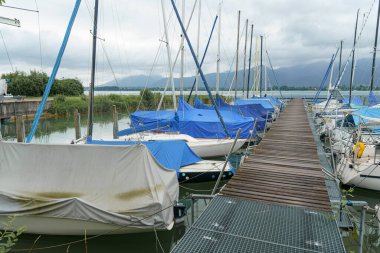 Birçok yelkenli, sakin sular ve bulutlu gökyüzünün altındaki uzak dağlarla çevrili mavi örtülü bir marinaya demirlemiştir..