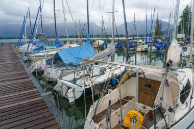 Birden fazla yelkenli, bazı gemileri koruyan mavi örtülü bir marinaya huzurlu bir şekilde yanaştı. Etrafındaki dağlar bulutlu bir gökyüzünün altında, sakin bir atmosfer yaratıyor..