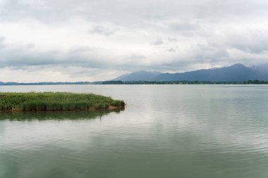 Sakin bir göl manzarası çevredeki dağları ve bulutlu gökyüzünü yansıtan sakin suları yansıtır. Yeşillik ve bataklık kıyı şeridinin kenarında. Sabah ışığı huzurlu atmosferi güçlendirir..
