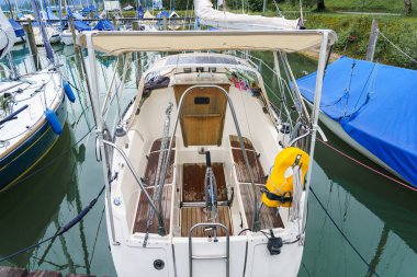 Bir yelkenli, sakin bir marinaya yanaşır. Ahşap güvertesi, sakin atmosferi yansıtır. Yakınlarda tekneler demirler ve sakin sular güneşli bir günde huzurlu bir manzara yaratır..