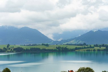 Bulutlar heybetli dağlar ve yeşilliklerle çevrili sakin bir göl üzerinde süzülüyor. Sahne, öğlen vakti suyun üzerindeki yansımalarla doğanın huzurlu atmosferini yakalıyor..