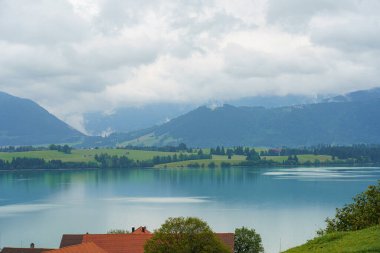 Gri bulutlar sakin bir gölün üzerinde yüzer, çevredeki dağları ve bitki örtüsünü yansıtır. Yakınlardaki evler tepenin yamacını işaretleyerek güzel bir kırsal alan oluşturuyor. Doğa güzelliğini sergiliyor.