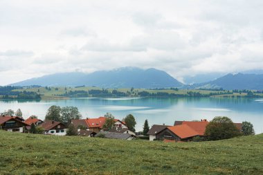 Sakin bir köy sakin bir göl kenarında, kırmızı çatıları olan büyüleyici evleri var. Dağlar kısmen bulutlarla kaplı. Sabahın erken saatlerinde sakin bir atmosfer yaratıyorlar..