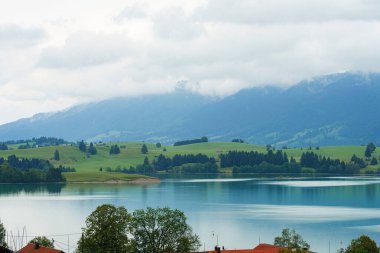 Sakin bir dağ gölü, etrafındaki yemyeşil tepelerin ve bulutlu bir gökyüzünün altındaki uzak tepelerin çarpıcı yansımalarını sergiliyor. Sakin su huzurlu bir atmosfer yaratır..