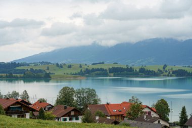 Bu huzurlu manzara, çevredeki dağları ve kıyı boyunca yuva yapmış büyüleyici evleri yansıtan sakin bir göle sahiptir. Bulutlar havada süzülür, yumuşak, sakin bir ortam ekler..