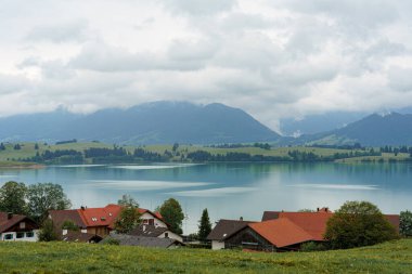 Gri bulutlar, etrafındaki dağları yansıtan sakin bir gölün üzerinde alçaktan süzülür. Yeşil tarlalar ve arka planda yuvarlanan tepeler tarafından vurgulanan tuhaf evler manzarayı benekler..