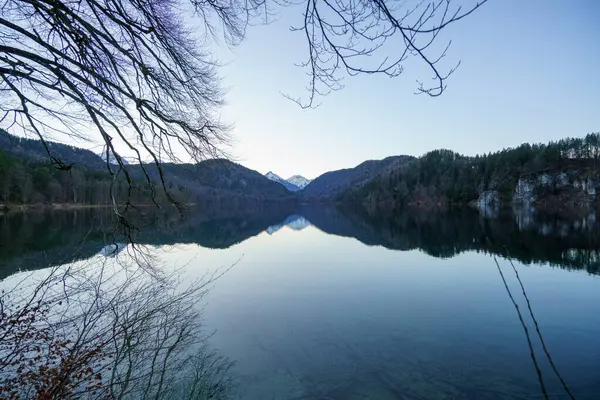 Gölün sakin suları çevredeki dağları yansıtıyor. Şafak vakti, huzurlu bir atmosfer yaratıyor. Çıplak dallar görüntüyü çerçeveler, sükuneti artırır.