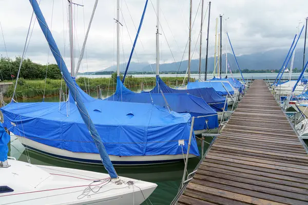 Mavi brandalarla kaplı birkaç yelkenli bir marinada hizaya sokuldu. Arkaplanda bulutlu gökyüzü belirirken, yemyeşil yemyeşil su ile sınır komşusudur. Sakin atmosfer huzurlu bir ortam olduğunu gösteriyor..