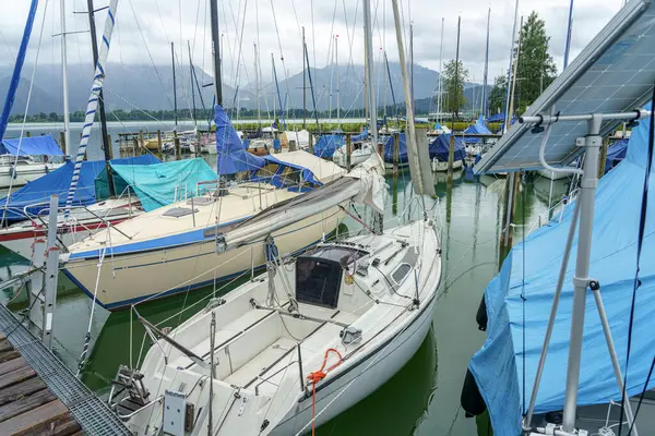 Yelkenliler ve yatlar yemyeşil ve dağlarla çevrili bir marinada demirli. Muşambalar, bulutlu bir günde hava korumasını sağlayan bazı gemileri kaplıyor..