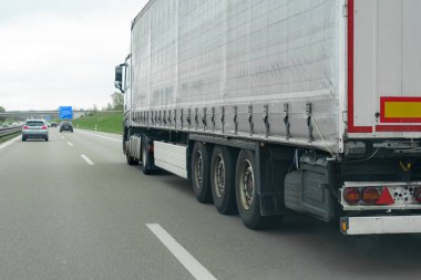 Çok şeritli bir otoyolda kargo taşıyan büyük bir kamyon, bulutlu bahar havasında daha küçük araçlarla birlikte. Yol kenarındaki yeşillik manzarayı güzelleştiriyor..