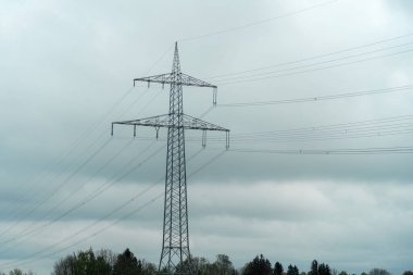 Kırsal bir alanda, bulutlu gökyüzü ve yeşil yapraklardan oluşan bir zemin üzerinde duran çelik şanzıman kulesi elektrik altyapısının önemini vurguluyor..