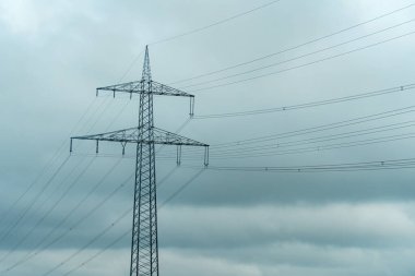 Uzun bir elektrik direği bulutlu bir gökyüzünün altında belirgin bir şekilde duruyor. Elektrik hatları yapıdan yayılarak modern yaşamda elektriğin önemini vurguluyor..