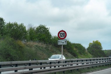 Saatte 120 km hızla giden bir hız limiti levhası 7 kilometre uzaklıktaki bir otoyolun yanında duruyor. Yakınındaki ağaçlarla bulutlu bir gökyüzünün altından arabalar geçiyor..