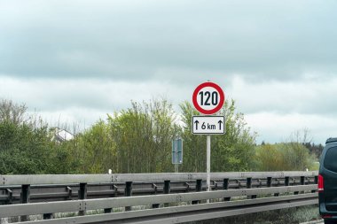 Bir hız limiti işareti maksimum 120 km / saat hız gösterirken, bir başka işaret hedefe 6 km uzaklık göstermektedir. Karayolu bulutlu gökyüzünün altında yemyeşil ağaçlarla kaplı..