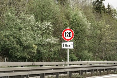 Saatte 120 km 'yi gösteren hız limiti levhası otoyolun kenarında belirgin bir şekilde gösteriliyor. Yeşiller yemyeşil ağaçlar ve yapraklar açık bir günde canlı bir zemin oluşturur..