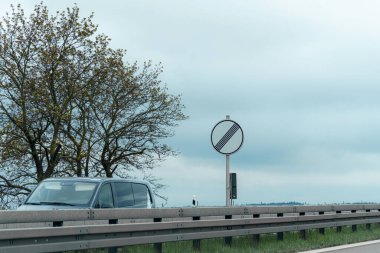 Otoyol kenarında kendine özgü bir yol işaretinin yanında bir araç park halinde. Çevre bulutlu gökyüzü ve yemyeşil arka planda sakin bir atmosfer yaratıyor..