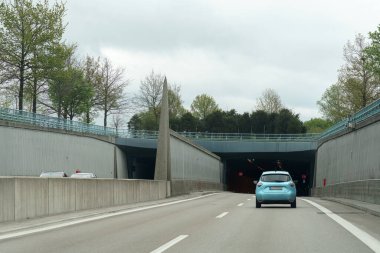Açık mavi bir araba gri gökyüzünün altındaki bir otoyol tüneline yaklaşıyor. Yol boyunca uzanan yemyeşil ağaçlar ilerideki tünel girişine geçişi vurguluyor..
