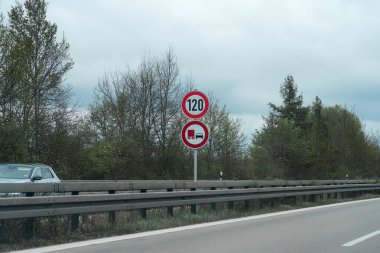 Saatte 120 kilometre hız sınırı gösteren bir otoyol hız limiti tabelası görülebilir. Yol ağaçlarla çevrili ve araçlar yakınlarda geziniyor. Bulutlu hava kasvetli bir ton ekler.