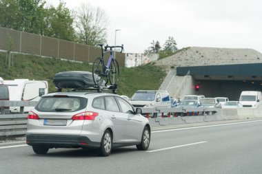 Gümüş renkli steyşın bir araba otobanda sürüyor, üstünde güvenli bir bisiklet var. Çevredeki trafik çeşitli araçları içeriyor, bu da yoğun bir işe gidiş-dönüş olduğunu gösteriyor.