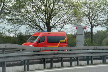 Trafik bariyerleri ve çevreleyen ağaçlarla bir otoyolun yakınında parlak kırmızı bir acil durum aracı konumlandırıldı. Bulutlu gökyüzü kırsal bölgedeki öğleden sonra koşullarını gösteriyor..