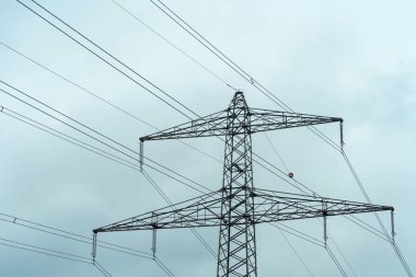 Bulutlu bir gökyüzüne yayılan sayısız elektrik hatları olan yüksek bir elektrik kulesi. Bu kurulum kırsal alandaki temel enerji aktarımını temsil ediyor..